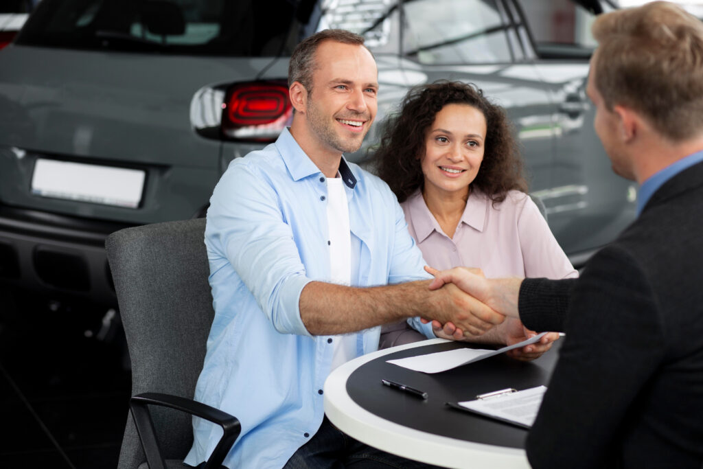 Smiling couple shaking hands with a car salesman at a dealership.