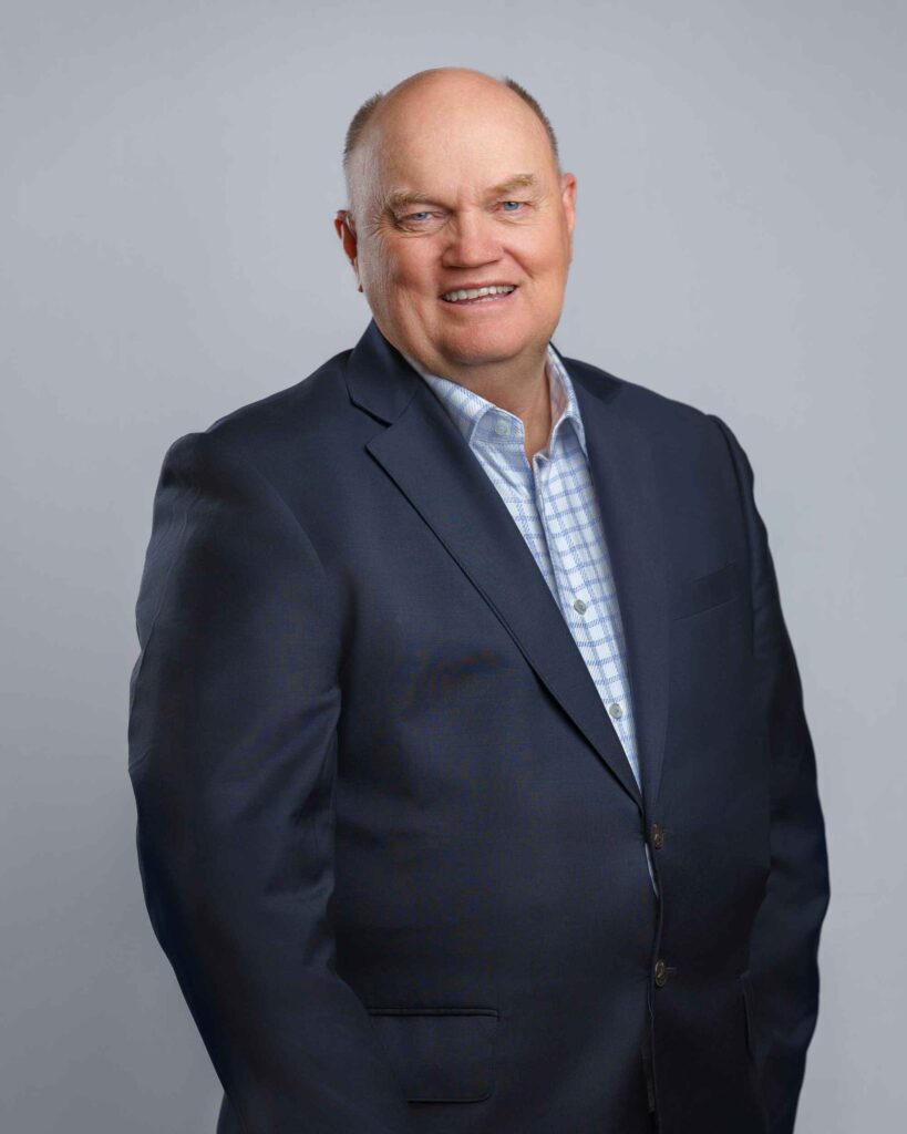 A middle-aged man in a dark blue suit and light blue plaid shirt smiling at the camera.