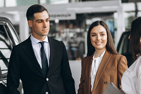 Two people in business attire standing near a vehicle, smiling at a third person.