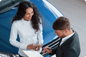 Woman and man in business attire discussing documents in front of a blue car.