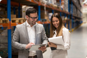 Man and woman in warehouse looking at tablet and paper.