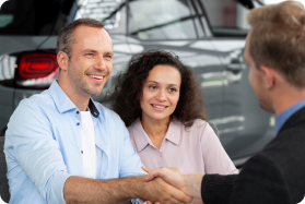 Smiling couple shaking hands with car salesman in front of vehicle.