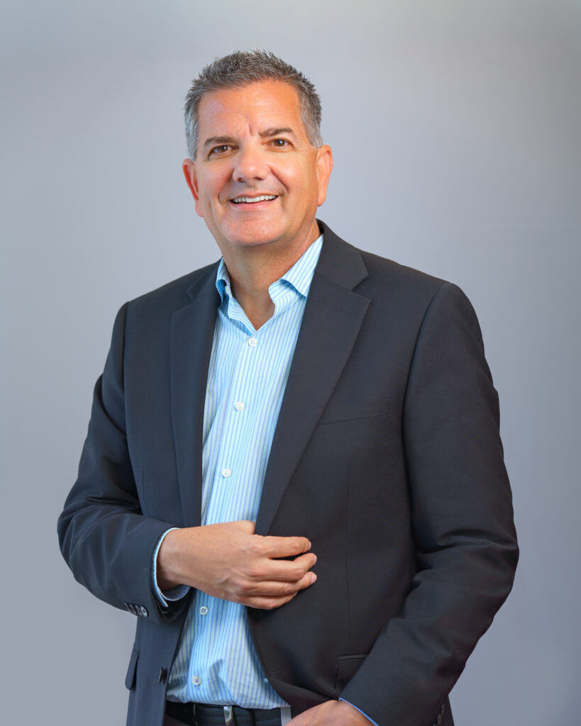 A middle-aged man in a dark suit jacket and light blue shirt smiling at the camera.