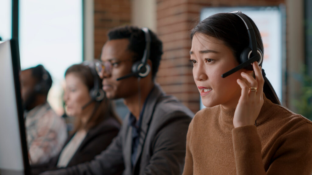 Woman wearing headset talking on phone with coworkers in background.