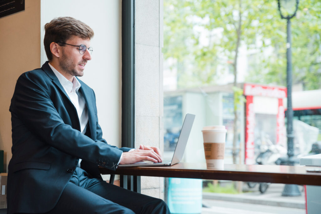 Man in blue suit working on laptop at table by window with coffee cup.