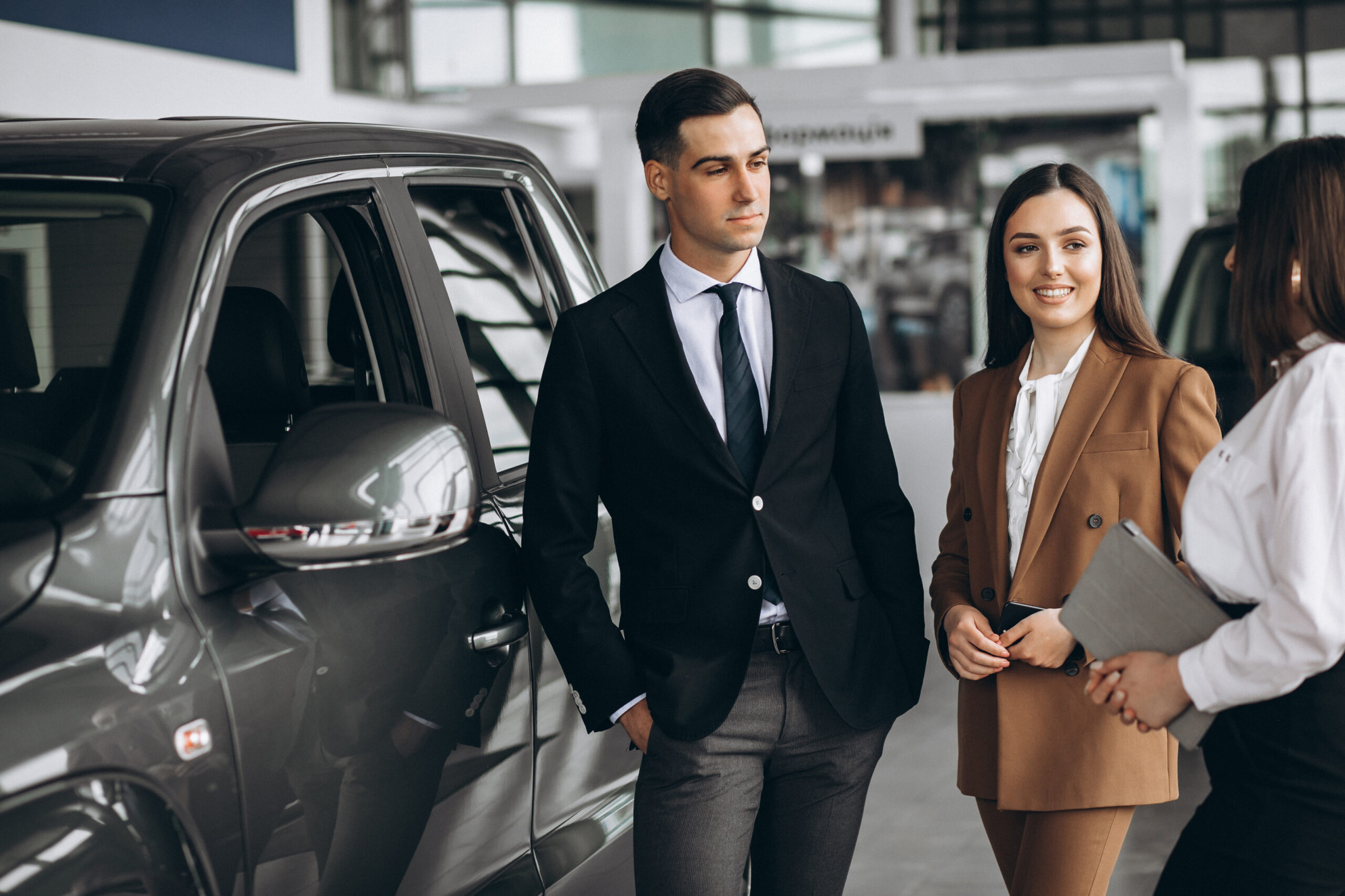 Man and woman in business attire standing next to a car talking to a woman with a clipboard.
